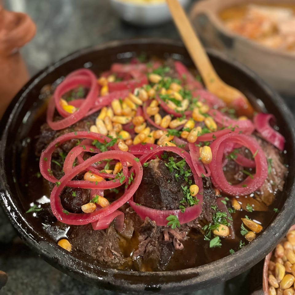 Braised meat served in a black dish, topped with pickled red onions, toasted nuts, and fresh herbs.