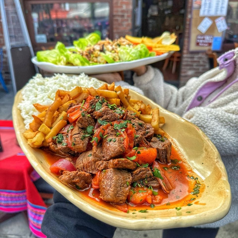 Large platter of beef stew with carrots, tomatoes, fries, and rice, held outside a brick restaurant alongside a salad plate.