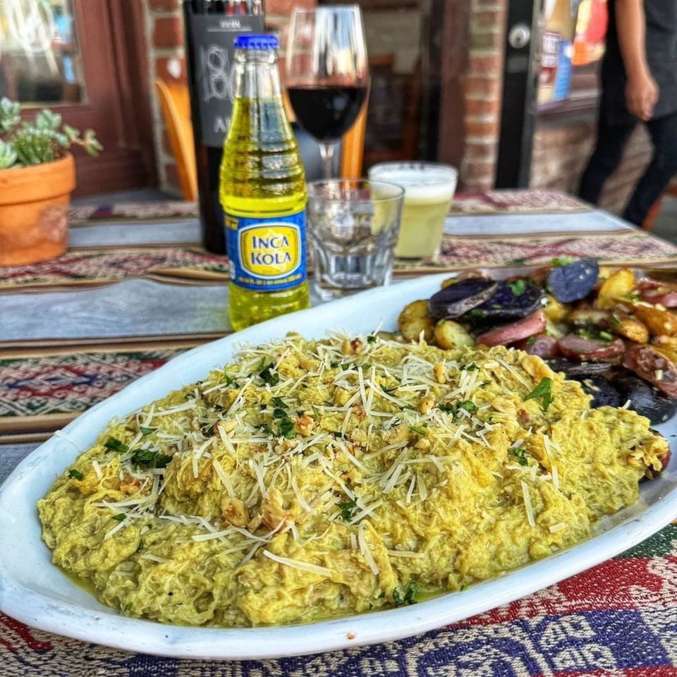 Large platter of creamy yellow dish topped with shredded cheese, herbs, and nuts, alongside roasted vegetables, wine, beer, and Inca Kola.